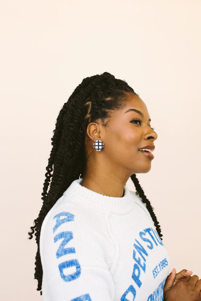 Woman wearing circular black and white plaid large stud earrings, handmade locally in Charlotte, NC (North Carolina), at a Plaza Midwood woman owned jewelry art and home decor gift shop. A can't miss thing to do - order a housemade vegan chai at The Chai Window to sip while you shop. 