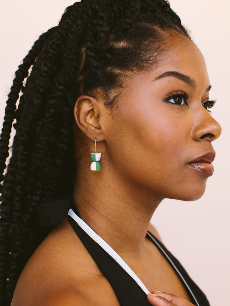 Woman wearing green, white, and gold hourglass shaped statement earrings, handmade locally in Charlotte, NC (North Carolina), at a Plaza Midwood woman owned jewelry art and home decor gift shop. A can't miss thing to do - order a housemade vegan chai at The Chai Window to sip while you shop. 