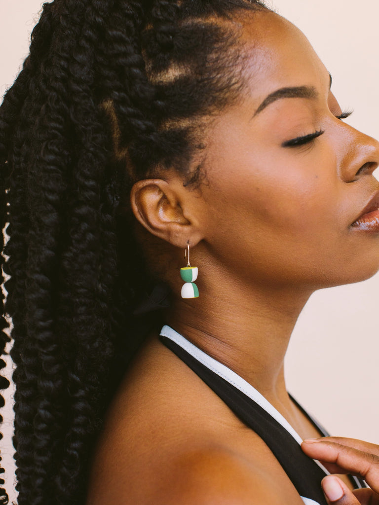 Woman wearing green, white, and gold hourglass statement earrings, handmade locally in Charlotte, NC (North Carolina), at a Plaza Midwood woman owned jewelry art and home decor gift shop. A can't miss thing to do - order a housemade vegan chai at The Chai Window to sip while you shop. 